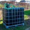 ibc-container-zwart-met-pomp-220V-1000L-vernieuwd-pallet-metaal-of-kunststof-zijaanzicht-met-uitgaande-slang-in-tuinomgeving.webp